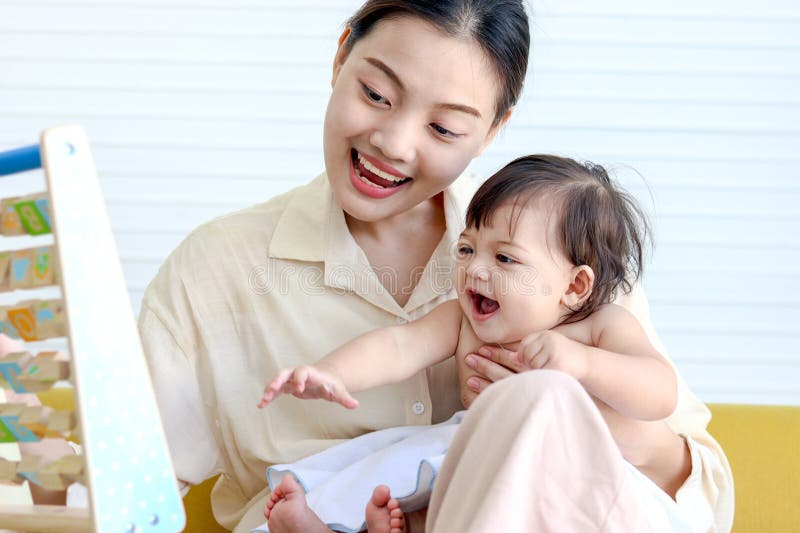 Happy smiling toddle baby sits on mother lap on yellow sofa in living room, little cute kid girl plays with developmental toys, mom plays with daughter child, love in family, childhood and motherhood. Happy smiling toddle baby sits on mother lap on yellow sofa in living room, little cute kid girl plays with developmental toys, mom plays with daughter child, love in family, childhood and motherhood