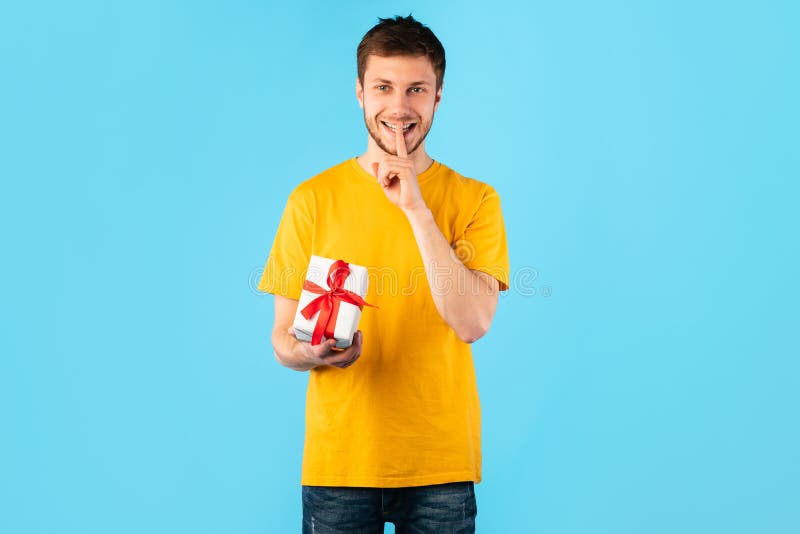 Surprise Gift. Portrait of smiling young guy holding wrapped present box and showing silence sign, isolated over blue studio background, greeting and celebrating special day or event. Surprise Gift. Portrait of smiling young guy holding wrapped present box and showing silence sign, isolated over blue studio background, greeting and celebrating special day or event