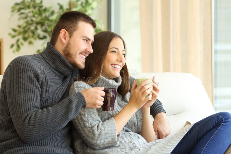 Affectionate couple wearing sweater holding hot coffee cups sitting on a sofa in the living room at home in winter. Affectionate couple wearing sweater holding hot coffee cups sitting on a sofa in the living room at home in winter