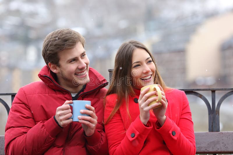 Happy couple drinking hot drinks looking at side in winter sitting on a bench in an urban park. Happy couple drinking hot drinks looking at side in winter sitting on a bench in an urban park