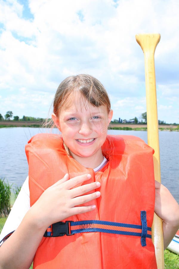 A happy and smiling girl in an orange life vest, holding a canoe paddle in front of a pond. A happy and smiling girl in an orange life vest, holding a canoe paddle in front of a pond.