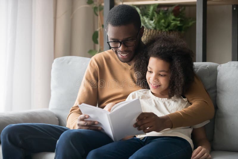 Happy family black father or baby sitter and kid daughter reading story book sitting on sofa together, african american child girl enjoying with daddy listening to fairy tale from dad at home. Happy family black father or baby sitter and kid daughter reading story book sitting on sofa together, african american child girl enjoying with daddy listening to fairy tale from dad at home