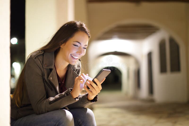 Gelukkig wijfje die een smartphone in de nacht in de straat met behulp van stock afbeelding Gelukkig wijfje die een smartphone in de nacht in de straat met behulp van stock afbeelding