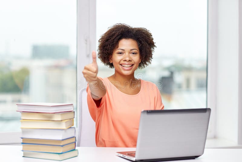 People, technology, gesture and education concept - happy african american young woman sitting at table with laptop computer and books showing thumbs up at home. People, technology, gesture and education concept - happy african american young woman sitting at table with laptop computer and books showing thumbs up at home