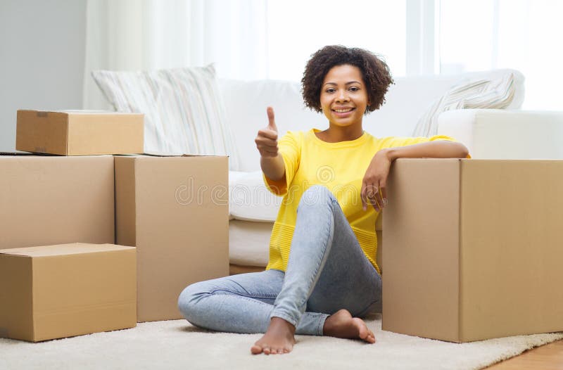 People, moving new place, gesture and repair concept - happy african american young woman with many cardboard boxes sitting on floor and showing thumbs up at home. People, moving new place, gesture and repair concept - happy african american young woman with many cardboard boxes sitting on floor and showing thumbs up at home