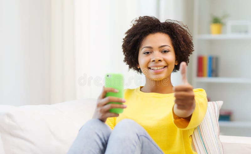 People, technology and leisure concept - happy african american young woman sitting on sofa with smartphone and showing thumbs up at home. People, technology and leisure concept - happy african american young woman sitting on sofa with smartphone and showing thumbs up at home