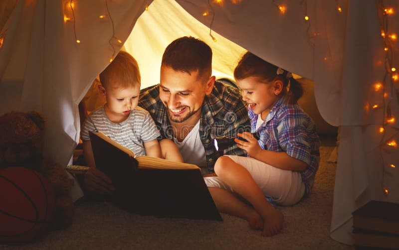 Happy family father and children reading a book with a flashlight in a tent at home. Happy family father and children reading a book with a flashlight in a tent at home