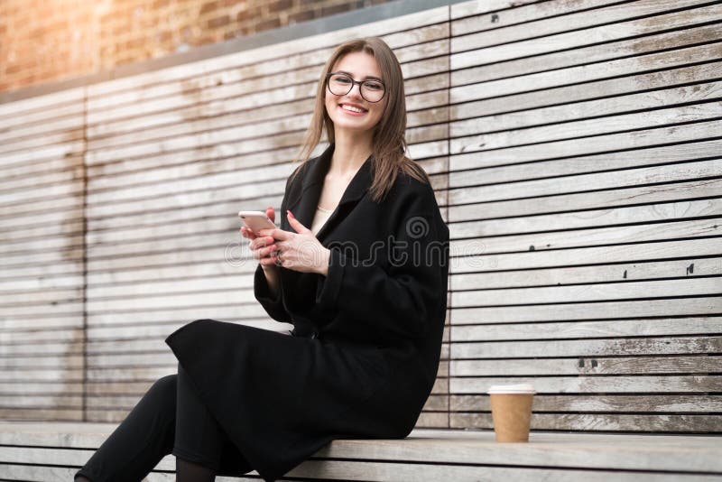 Gelukkige het glimlachen vrouwenzitting in openlucht en het texting op haar telefoon het drinken koffie royalty-vrije stock afbeeldingen Gelukkige het glimlachen vrouwenzitting in openlucht en het texting op haar telefoon het drinken koffie royalty-vrije stock afbeeldingen