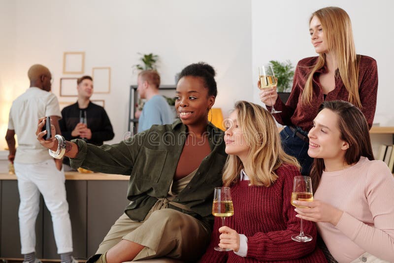 Happy young intercultural women with flutes of champagne making selfie while cheering up and enjoying home party against their boyfriends. Happy young intercultural women with flutes of champagne making selfie while cheering up and enjoying home party against their boyfriends