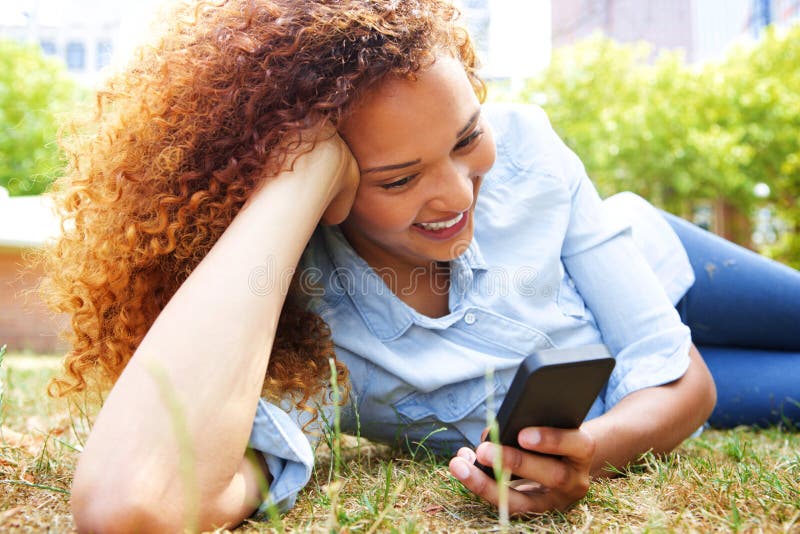 Gelukkige jonge vrouw die in gras bij het park liggen en mobiele telefoon bekijken royalty-vrije stock foto Gelukkige jonge vrouw die in gras bij het park liggen en mobiele telefoon bekijken royalty-vrije stock foto