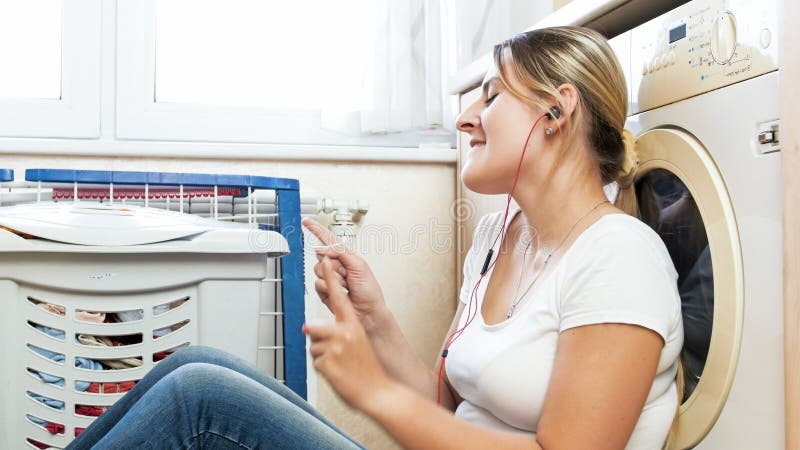 Gelukkige jonge vrouw het luisteren muziek met oortelefoons terwijl het doen van huishoudelijk werk bij wasserijruimte royalty-vrije stock foto Gelukkige jonge vrouw het luisteren muziek met oortelefoons terwijl het doen van huishoudelijk werk bij wasserijruimte royalty-vrije stock foto