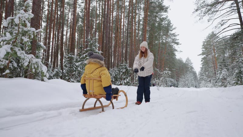 Gelukkige jonge vrouw loopt met haar kleine kind in het winterwoud . zoon zit in houten slee