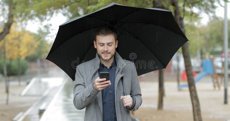 Gelukkige mens die telefoon controleren die onder de regen lopen