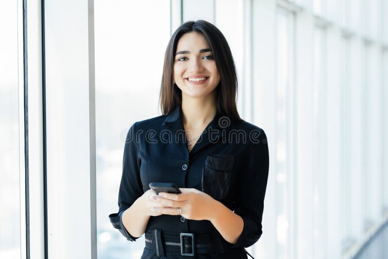 Gelukkige onderneemster die smartphone in bureau gebruiken dichtbij venster stock fotografie Gelukkige onderneemster die smartphone in bureau gebruiken dichtbij venster stock fotografie