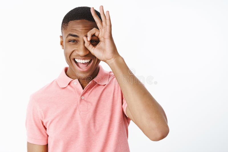 Happy uplifted and charismatic attractive young african american man smiling and laughing joyfully showing zero or okay gesture over eye looking through it happily over white background. Emotions and advertising concept. Happy uplifted and charismatic attractive young african american man smiling and laughing joyfully showing zero or okay gesture over eye looking through it happily over white background. Emotions and advertising concept