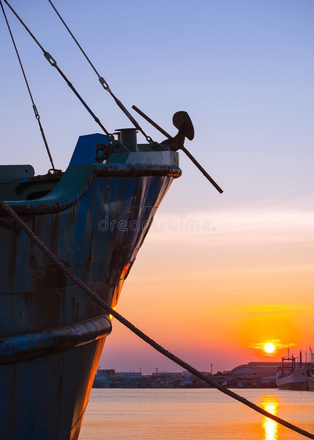 Silhouette forward side of fishing boat moored at harbor with mooring rope against sunset sky background in evening time