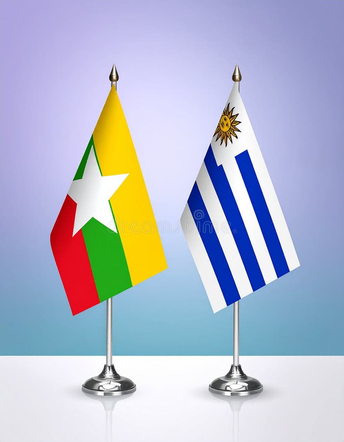 Two table flags on stands are displayed against a gradient blue background. The flag on the left is Myanmar's, with horizontal stripes of yellow, green, and red and a large white star in the center. The flag on the right is Uruguay's, featuring nine alternating white and blue stripes with a white canton and a golden Sun of May. Both flags are mounted on small black stands with a reflective surface below. Myanmar and Uruguay flags the relationship with the table, gradient background. Vector. Al Generated. Canton flags illustrations