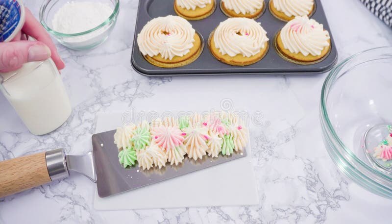 A kitchen scene with cookies in a muffin tin topped with swirled white frosting and sprinkled pink decorations. A spatula holds piped dollops of frosting in light pink, white, and green. A small glass bowl contains white sprinkles, while a person's hand holds a small glass bottle. The surface is a white marble countertop, suggesting a baking process. Frosting spatula illustrations