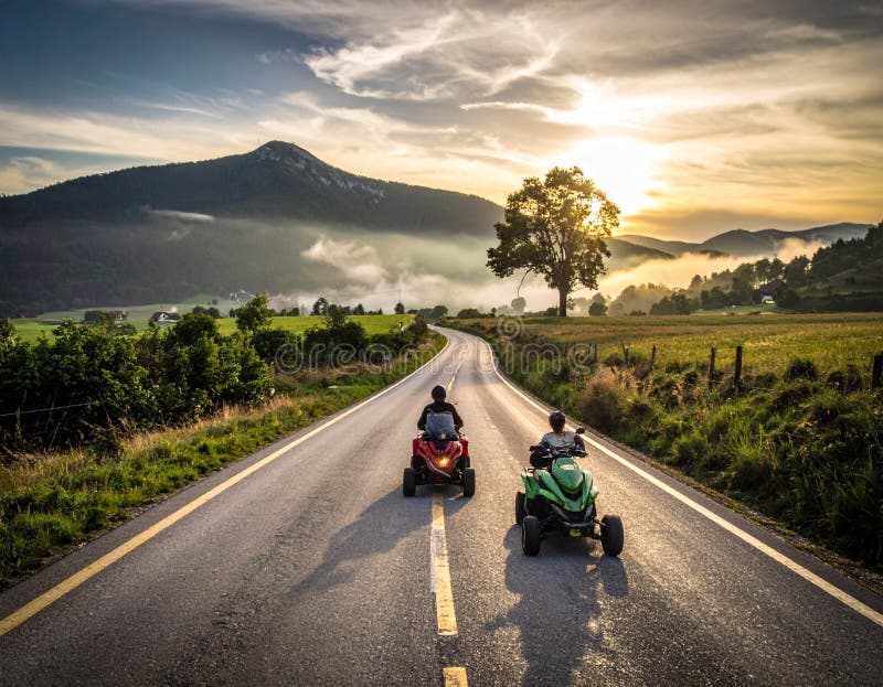 Two people ride quad bikes on a rural road, backed by a stunning landscape of mountains stock illustration