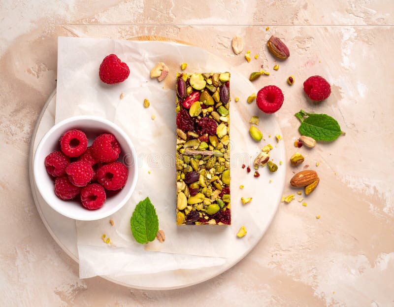 A rectangular fruit and nut bar is placed on a brown ceramic plate. The bar is topped with vibrant red raspberries (Rubus idaeus) and green pistachios (Pistacia vera). Surrounding the plate on a dark textured surface are additional raspberries, almonds (Prunus dulcis), and pistachios. A small bowl holds more pistachios and raspberries. The arrangement emphasizes the natural colors and textures of the ingredients, creating a visually appealing and appetizing display. Raspberry & Pistachio Protein Bar with fruit full bird's eye shot. Al Generated. Ceramic birds illustrations