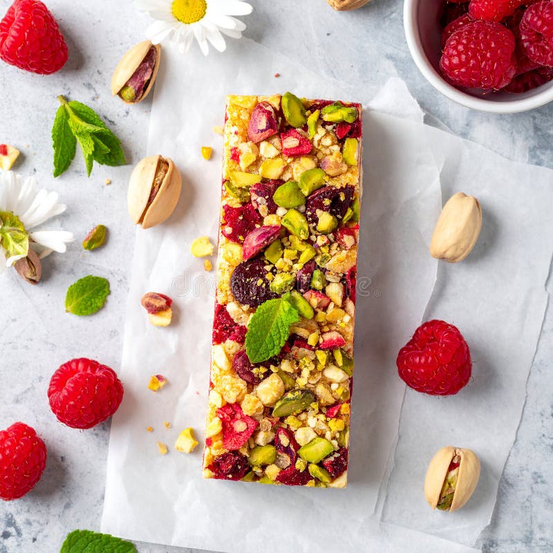 A rectangular fruit and nut bar is placed on a brown ceramic plate. The bar is topped with vibrant red raspberries (Rubus idaeus) and green pistachios (Pistacia vera). Surrounding the plate on a dark textured surface are additional raspberries, almonds (Prunus dulcis), and pistachios. A small bowl holds more pistachios and raspberries. The arrangement emphasizes the natural colors and textures of the ingredients, creating a visually appealing and appetizing display. Raspberry & Pistachio Protein Bar with fruit full bird's eye shot. Al Generated. Ceramic birds illustrations