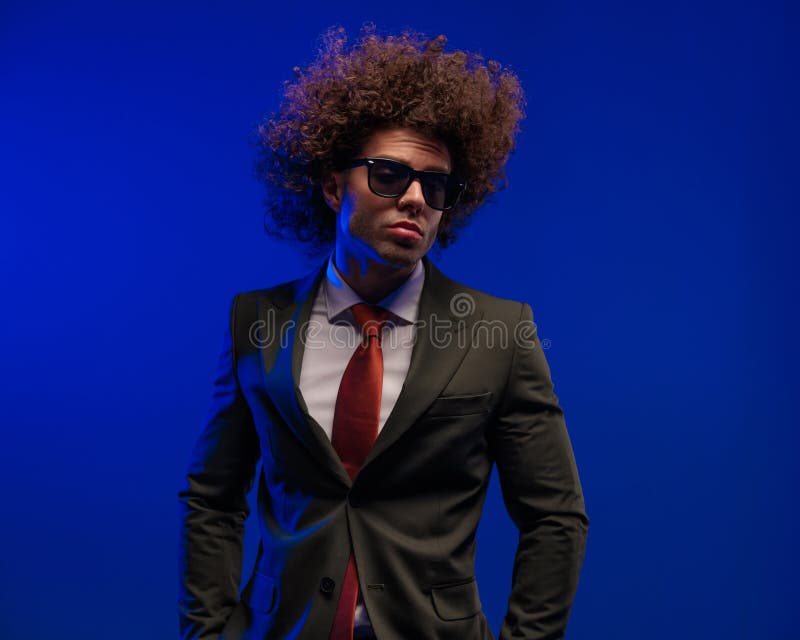 Genial para un hombre de negocios con gafas de sol agarrando las manos en los bolsillos. Genial para un hombre de negocios con traje negro con gafas de sol cogiendo la mano en los bolsillos y posando frente al fondo azul