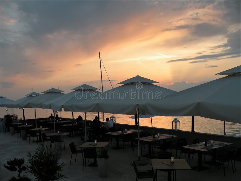 Gente en la cena, tablas debajo de los paraguas blancos Restaurante por el mar Puesta del sol La escena mediterránea del día de f fotos de archivo Gente en la cena, tablas debajo de los paraguas blancos Restaurante por el mar Puesta del sol La escena mediterránea del día de f fotos de archivo