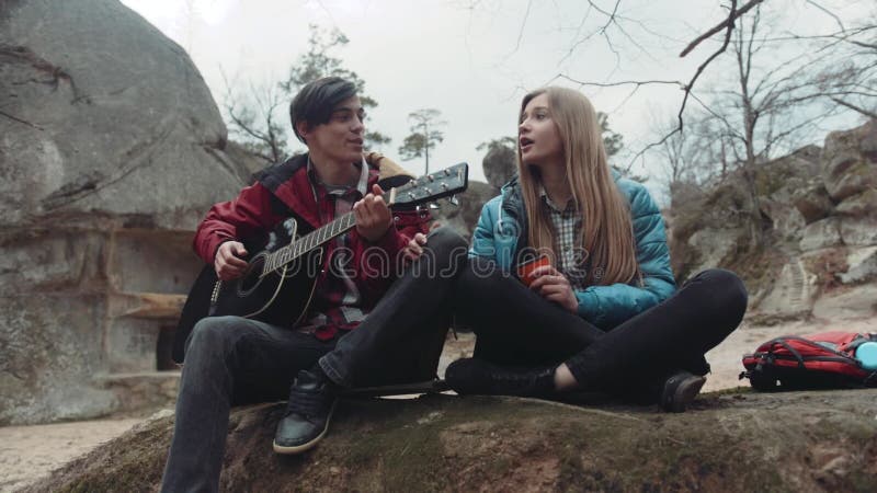 Gente joven que se sienta en la roca y que canta una canción, hombre joven que toca la guitarra, y consumición rubia encantadora