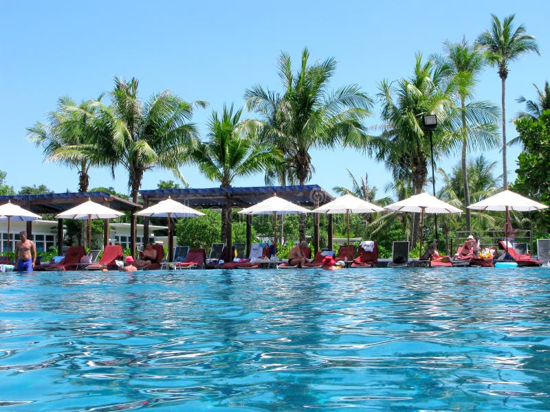 Asia, Thailand, Phuket island. People relax at swimming pool of hotel. Tropical climate. High palm trees, beach umbrellas and sunbeds. Turquoise reflection of sky on water surface in pool. Asia, Thailand, Phuket island. People relax at swimming pool of hotel. Tropical climate. High palm trees, beach umbrellas and sunbeds. Turquoise reflection of sky on water surface in pool.