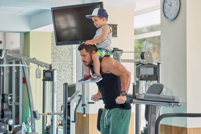 Geschikte sportman opleiding op gymnastiek` s simulator die weinig zoon op hals houden royalty-vrije stock fotografie Geschikte sportman opleiding op gymnastiek` s simulator die weinig zoon op hals houden royalty-vrije stock fotografie