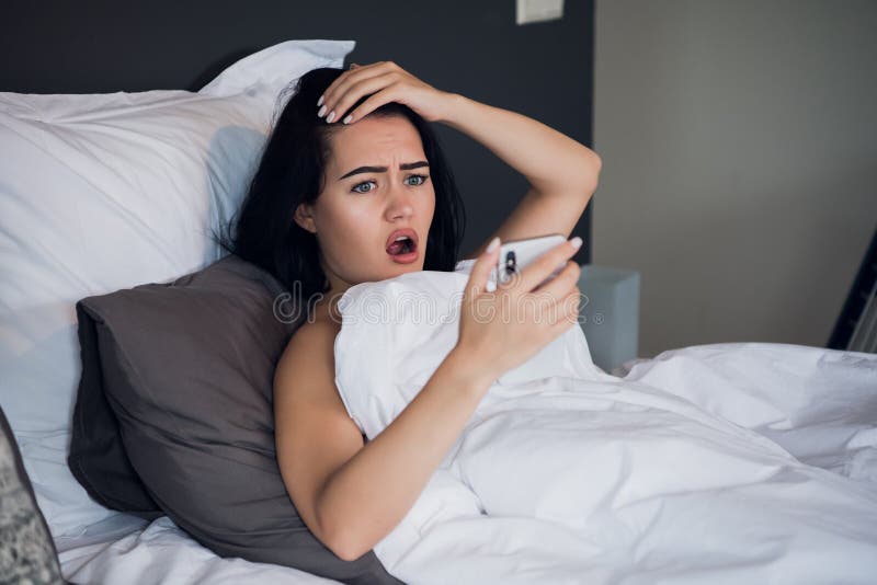 Geschokte jonge vrouw die mobiele telefoon in bed thuis bekijken royalty-vrije stock foto's Geschokte jonge vrouw die mobiele telefoon in bed thuis bekijken royalty-vrije stock foto's