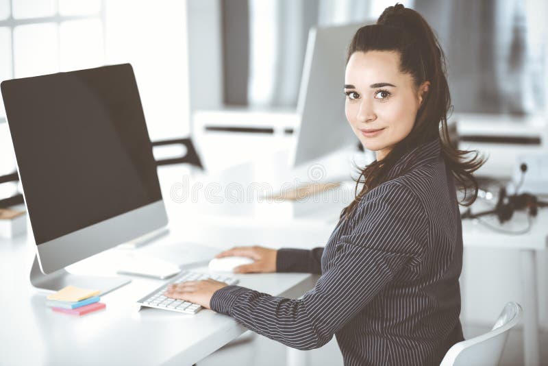 Geschäftsfrau mithilfe des Computers am Arbeitsplatz im Büro. brunette Sekretär oder weiblicher Anwalt, lächelt und sieht glücklic. Generalsekretär oder weiblicher Anwalt, welche die Kamera und das glückliche Lächeln sehen. Funktionieren für Freude und Erfolg.