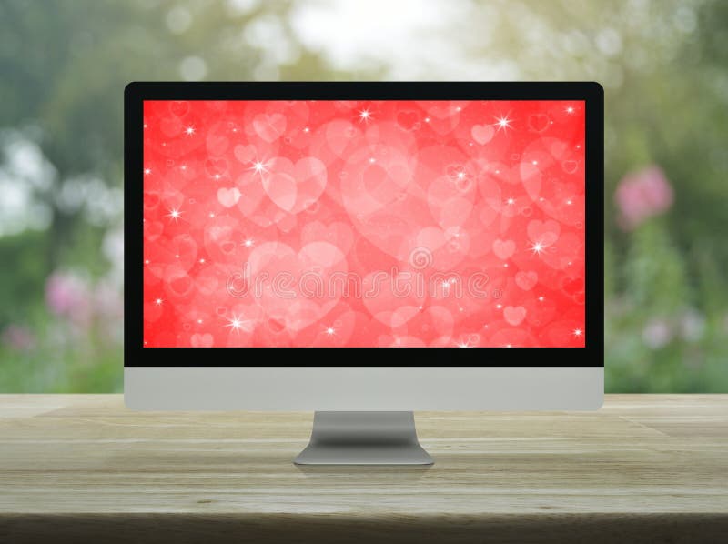 Geschäftsinternet-Datierungson-line-Valentinsgrußtageskonzept. Moderner Computertischplattenmonitor mit rotem Liebesherzschirm auf Holztisch über rosa Blume der verschwommener Fleck und Baum Parkgeschäftsinternet-Datierungsim on-line-Valentinsgrußtageskonzept