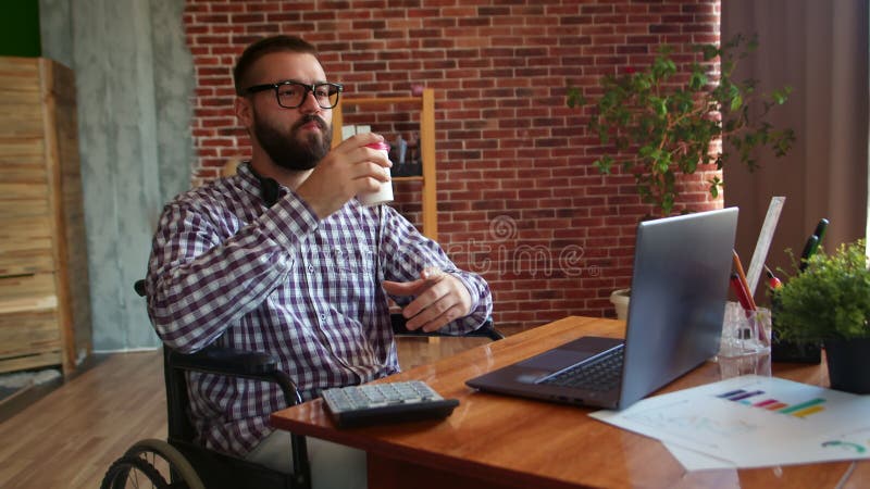 Geschäftsmann mit Brille im Rollstuhl trinkt Kaffee an seinem Schreibtisch. Hipster behindert arbeiten im Büro nahm Pause