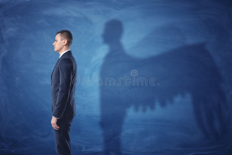 Geschäftsmann steht im Profil und wirft einen Schatten von Engelsflügeln auf blauem Tafelhintergrund lizenzfreies stockbild Geschäftsmann steht im Profil und wirft einen Schatten von Engelsflügeln auf blauem Tafelhintergrund lizenzfreies stockbild