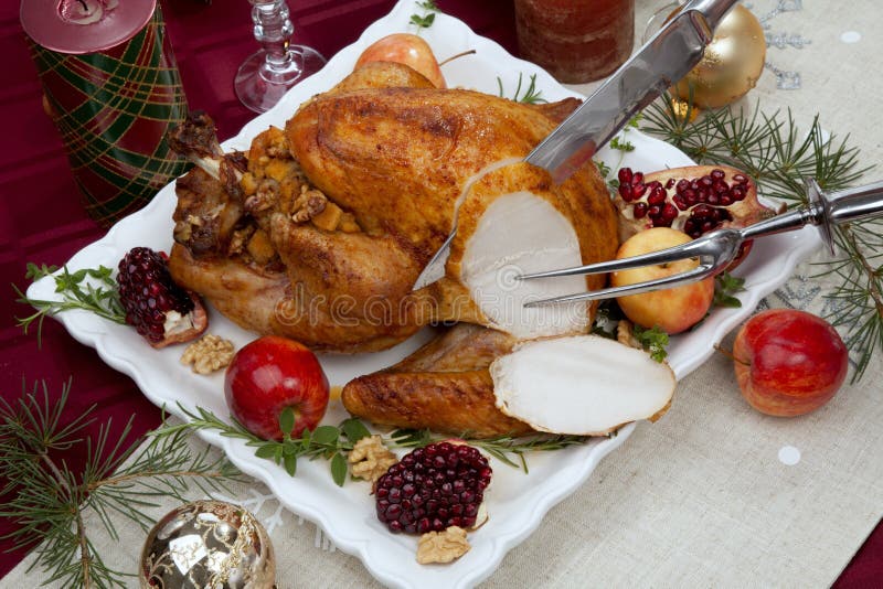 Carving pomegranate glazed roasted turkey on a tray garnished with fresh pomegranates, apples, herbs, and walnuts on Christmas-decorated table with candles, ornaments, fir twigs and flutes of champagne. Carving pomegranate glazed roasted turkey on a tray garnished with fresh pomegranates, apples, herbs, and walnuts on Christmas-decorated table with candles, ornaments, fir twigs and flutes of champagne