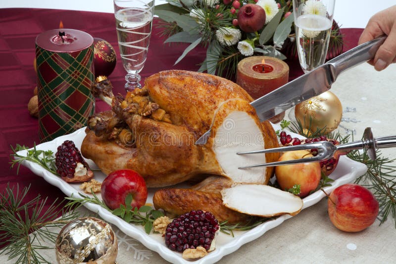 Carving pomegranate glazed roasted turkey on a tray garnished with fresh pomegranates, apples, herbs, and walnuts on Christmas-decorated table with candles, ornaments, fir twigs and flutes of champagne. Carving pomegranate glazed roasted turkey on a tray garnished with fresh pomegranates, apples, herbs, and walnuts on Christmas-decorated table with candles, ornaments, fir twigs and flutes of champagne