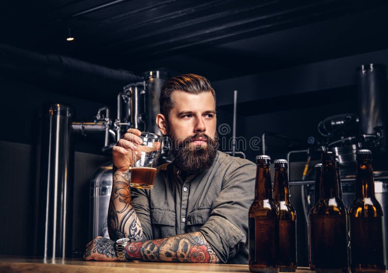 Getatoeeerd hipster mannetje met modieuze baard en haar het drinken bierzitting bij de barteller in de indiebrouwerij royalty-vrije stock fotografie Getatoeeerd hipster mannetje met modieuze baard en haar het drinken bierzitting bij de barteller in de indiebrouwerij royalty-vrije stock fotografie