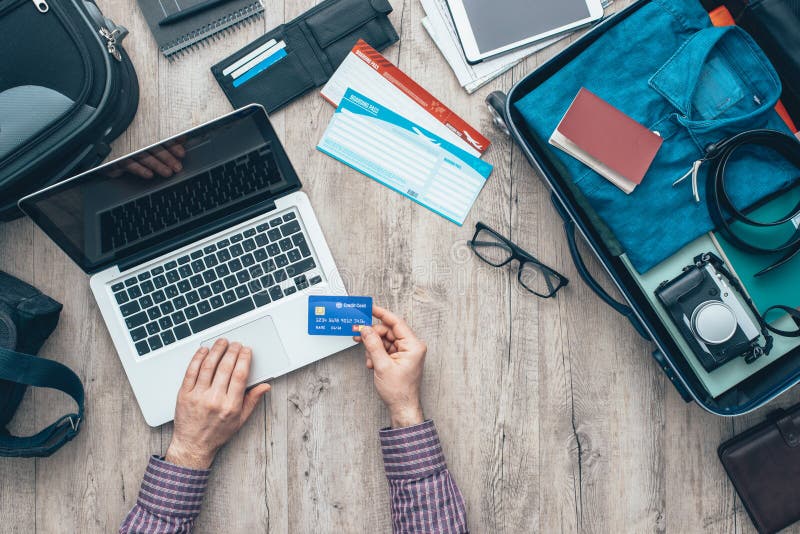 Traveler getting ready for a trip, he is packing his bag and booking a flight online using a credit card and a laptop, flat lay