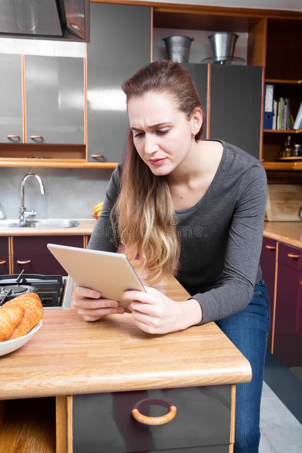 Geërgerde jonge vrouw die in lezing op tablet in keuken fronsen royalty-vrije stock foto Geërgerde jonge vrouw die in lezing op tablet in keuken fronsen royalty-vrije stock foto