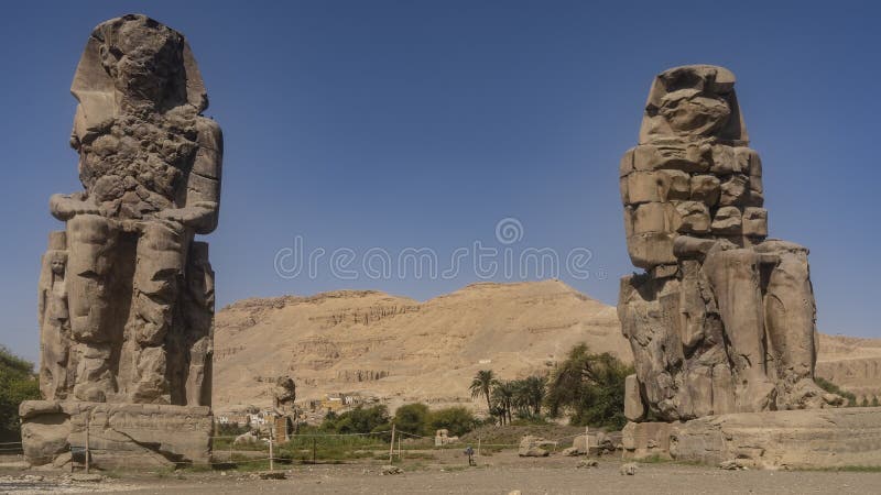 Giant sculptures of the colossi of Memnon against a blue sky and a sand dune.