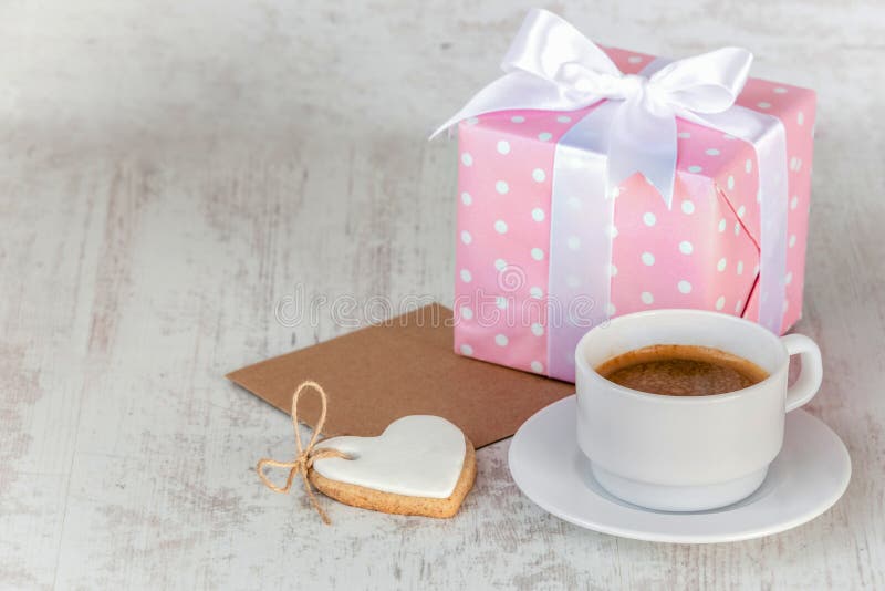 Gift box wrapped in pink dotted paper, heart shaped love cookie, a cup of coffee and an empty kraft card over a white wood