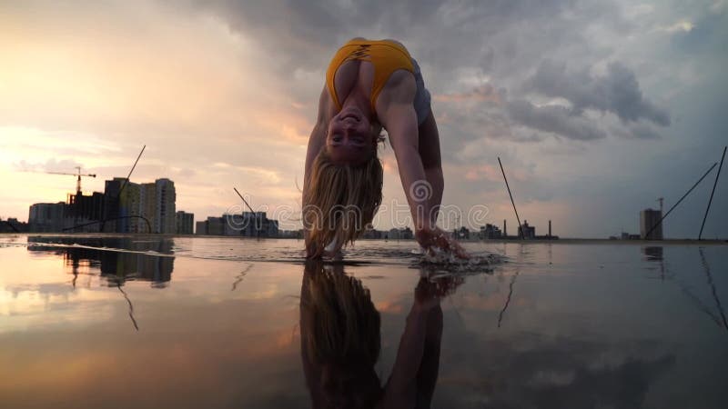 Gimnasta femenina flexible rubia va cabeza abajo en el puente con reflexión en el agua sobre fondo de paisaje urbano
