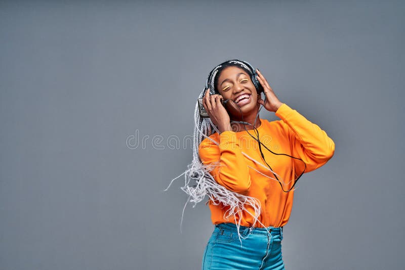 Gioiosa donna afro-americana vestita casualmente danzando e ascoltando musica con piacere con cuffie bianche isolate in grigio fotografia stock