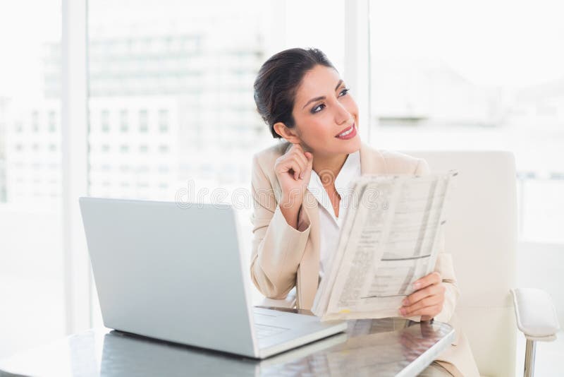 Giornale sorridente della tenuta della donna di affari mentre lavorando al computer portatile fotografia stock Giornale sorridente della tenuta della donna di affari mentre lavorando al computer portatile fotografia stock