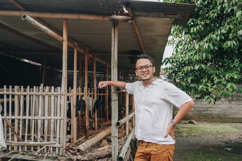 Giovane asiatico eccitato di fronte alla gabbia tradizionale fatta di legno e bambù nella zona rurale indonesiana con capra all'in fotografia stock