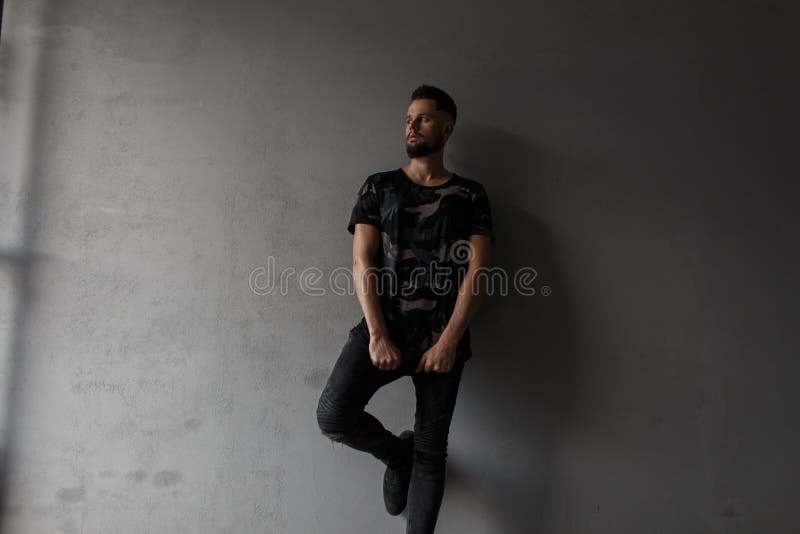 Giovane attraente grazioso con un'acconciatura alla moda e una barba brutale in una camicia alla moda militare, jeans neri immagine stock