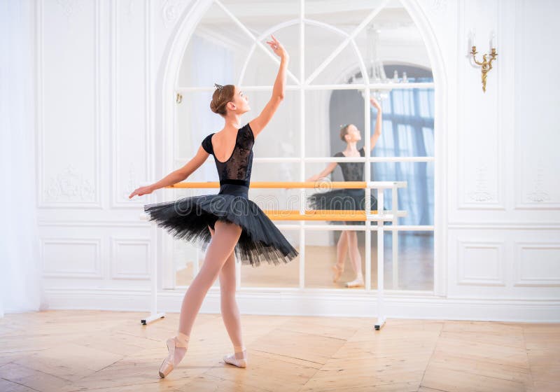 Giovane ballerina in un tutu nero in posa graziosa sulle scarpe a punta in una grande sala luminosa di fronte a uno specchio