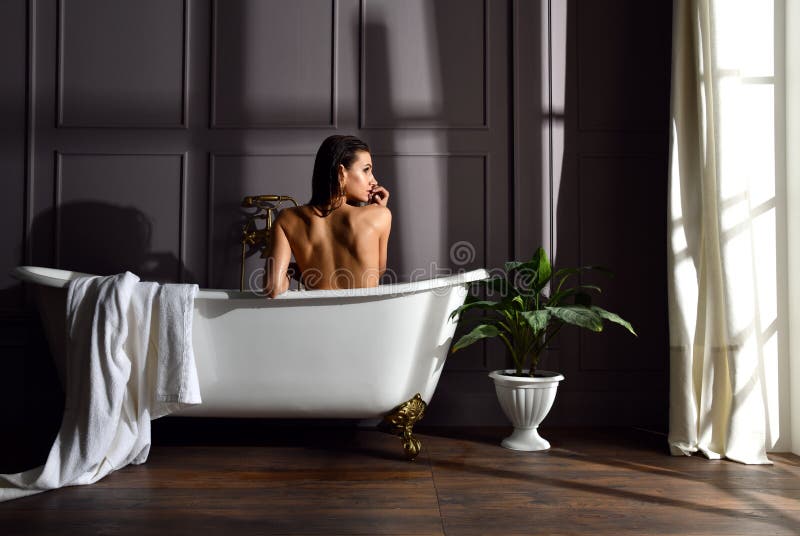 Giovane bella donna che si siede nel bagno vicino al bagno costoso della vasca che esamina l'angolo sul buio fotografia stock Giovane bella donna che si siede nel bagno vicino al bagno costoso della vasca che esamina l'angolo sul buio fotografia stock
