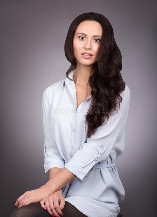 Giovane bella donna con capelli scuri lunghi e la camicia blu che posano su grigio immagine stock Giovane bella donna con capelli scuri lunghi e la camicia blu che posano su grigio immagine stock